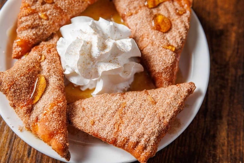 Four triangular pastries dusted with cinnamon sugar, drizzled with honey, and topped with whipped cream.
