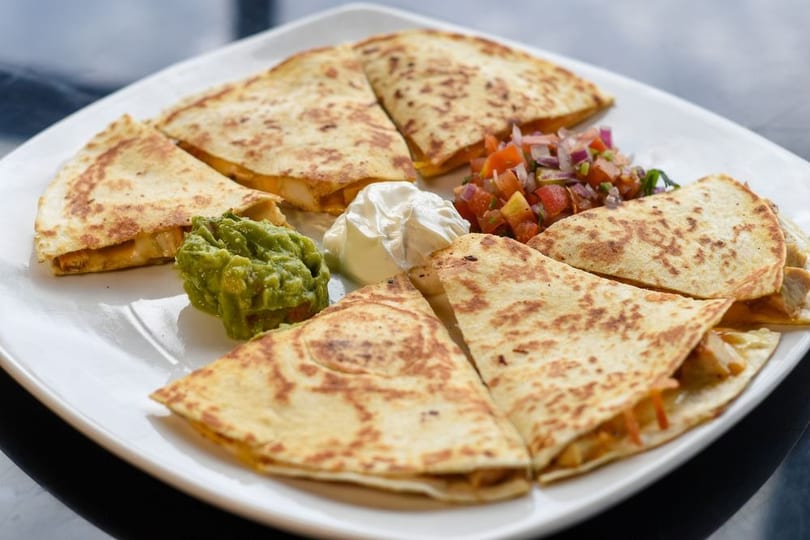 A white plate with six quesadilla wedges, guacamole, sour cream, and pico de gallo.
