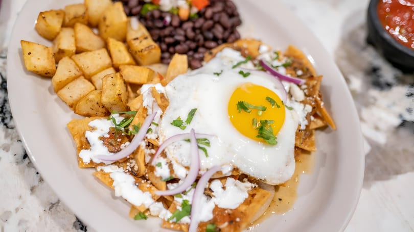 Chilaquiles with a fried egg, sour cream, red onions, cilantro, and a side of potatoes and black beans.