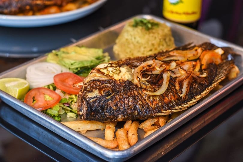 Whole fried fish with onions, rice, fries, avocado, salad, and lime on a metal tray.