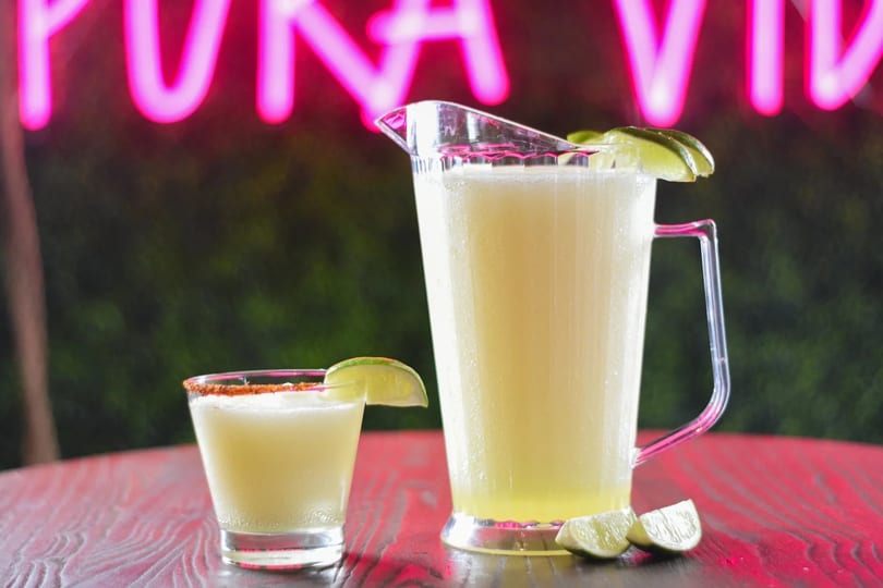A margarita glass and pitcher with lime slices, against a neon pink sign.