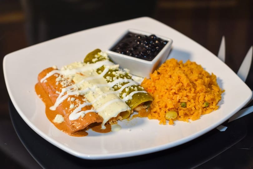 Enchiladas with red and green sauce, rice, and black beans on a white plate.