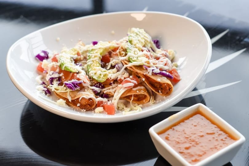 Crispy taquitos topped with avocado, tomatoes, shredded cabbage, cheese, and crema, with a side of salsa.