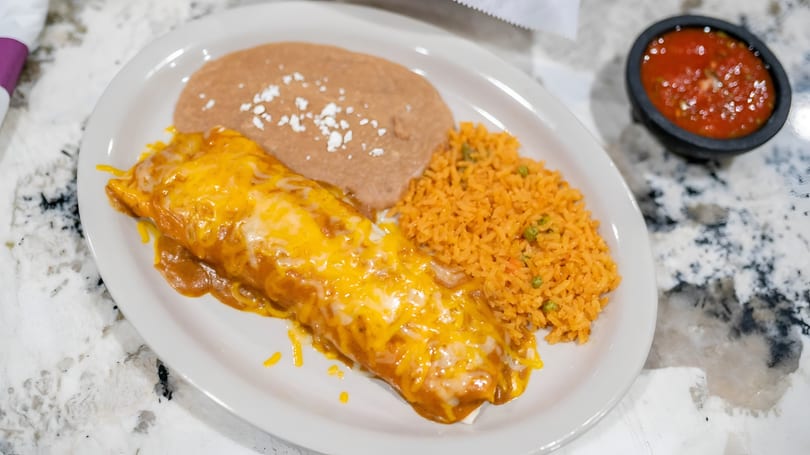 A cheesy burrito with refried beans and Mexican rice on a white plate, with a side of salsa.