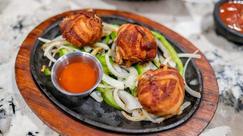 Three bacon-wrapped appetizers with onions and peppers on a sizzling platter with dipping sauce.