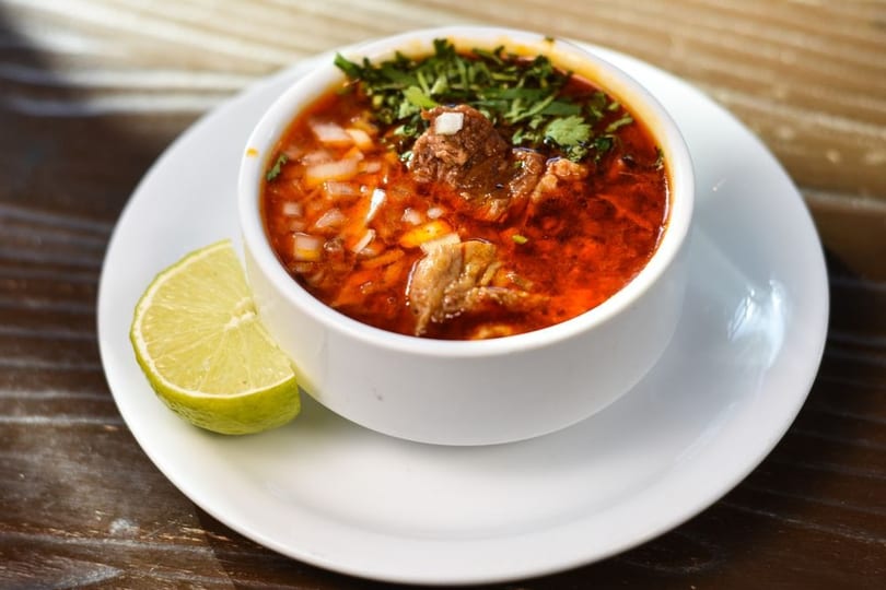 Hearty red soup with meat, onions, and cilantro in a white bowl with a lime wedge.