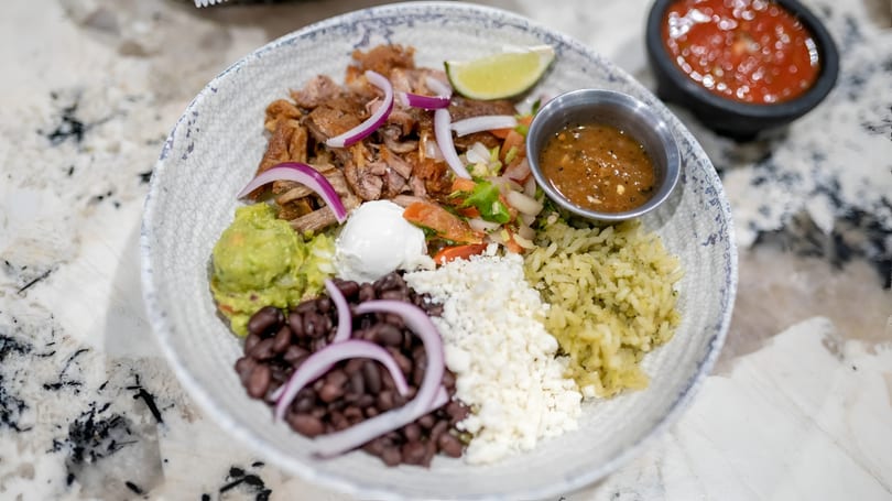 A colorful bowl of carnitas, rice, black beans, guacamole, cheese, and salsa on a marble counter.