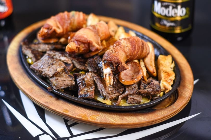 Sizzling platter of steak, bacon-wrapped shrimp, sausage, and fries, with a Modelo beer in the background.
