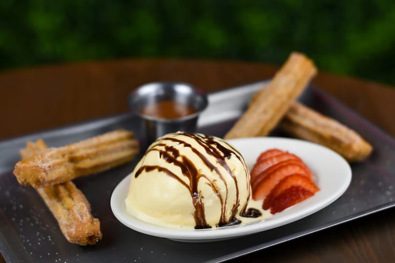 Vanilla ice cream with chocolate drizzle, sliced strawberries, churros, and caramel sauce on a tray.