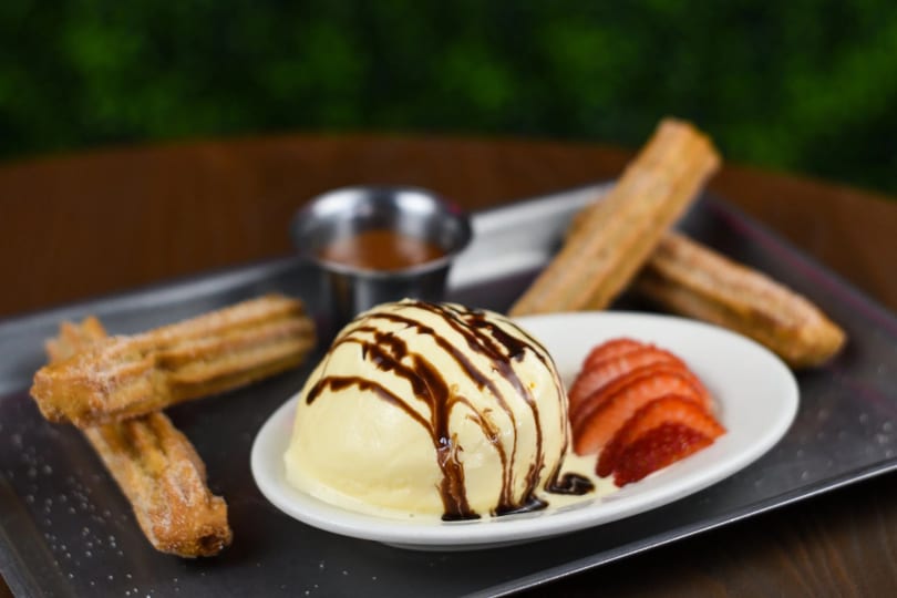Churros with vanilla ice cream drizzled with chocolate, sliced strawberries, and a side of caramel.