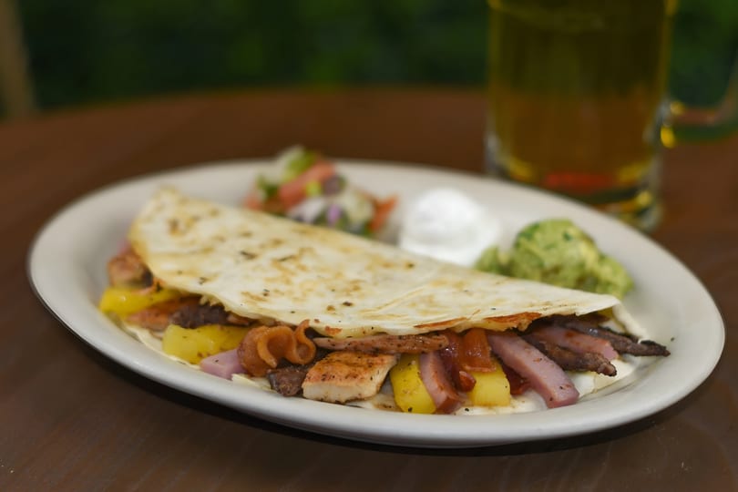 A quesadilla filled with chicken, ham, pineapple, and bacon, served with guacamole, sour cream, and salsa.