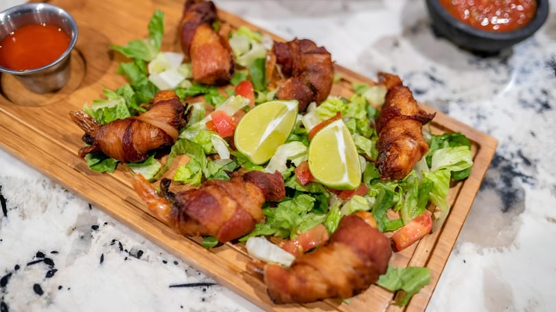 Bacon-wrapped shrimp on a bed of lettuce and diced tomatoes with lime wedges and a side of sauce.