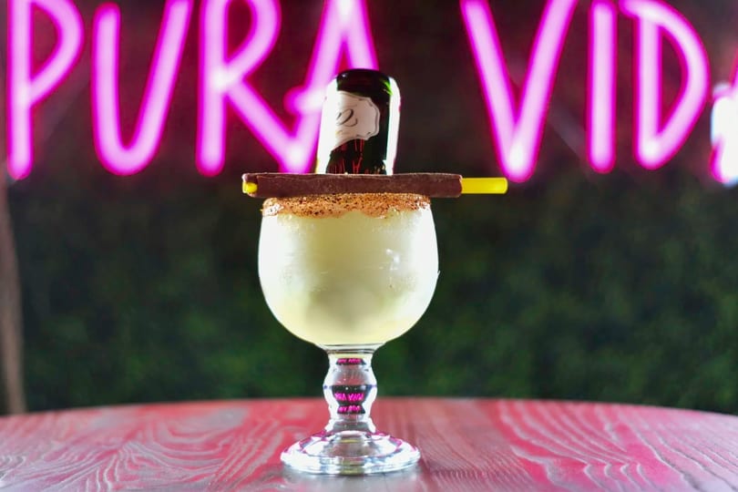 Frozen margarita with a mini beer bottle, chocolate stick, and yellow straw against a neon sign.