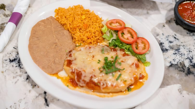 A white plate with a cheesy enchilada, refried beans, orange rice, and a side salad.