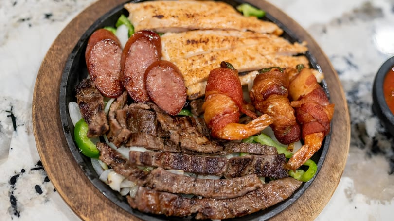 Sizzling fajita platter with steak, chicken, sausage, bacon-wrapped shrimp, onions, and peppers.