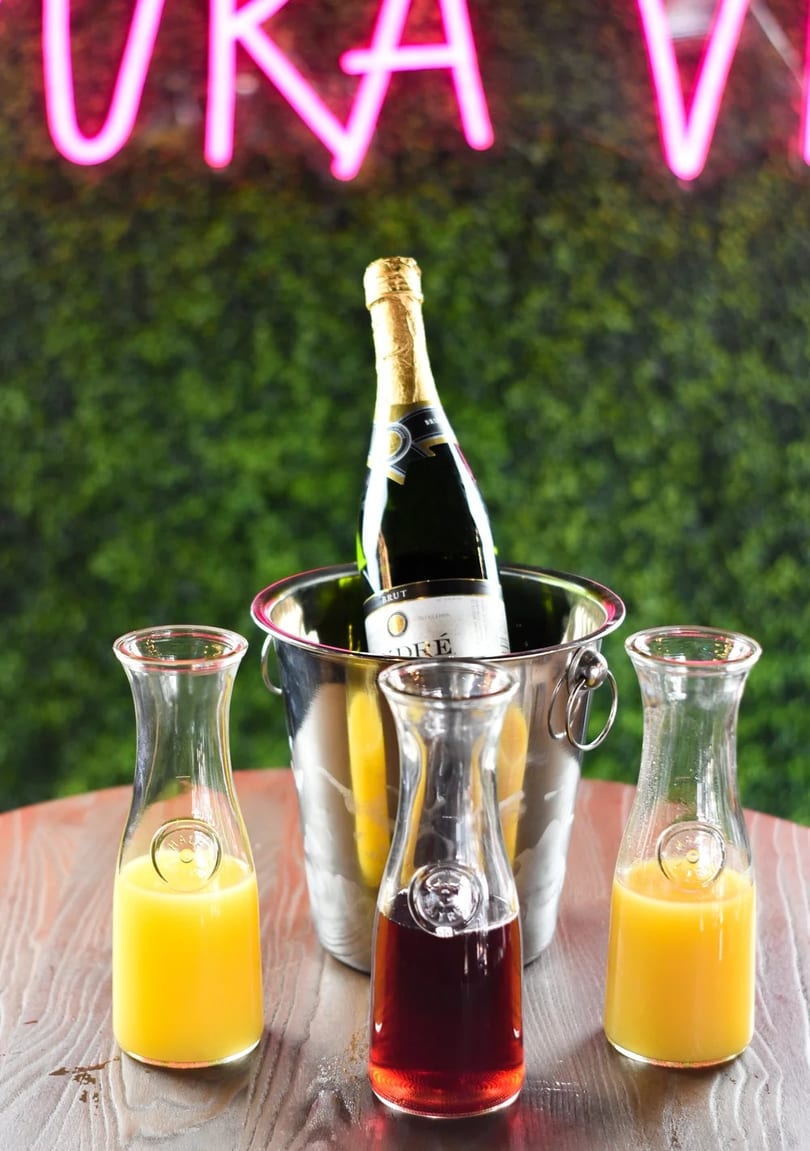 Champagne bottle in an ice bucket with three carafes of juice on a wooden table.